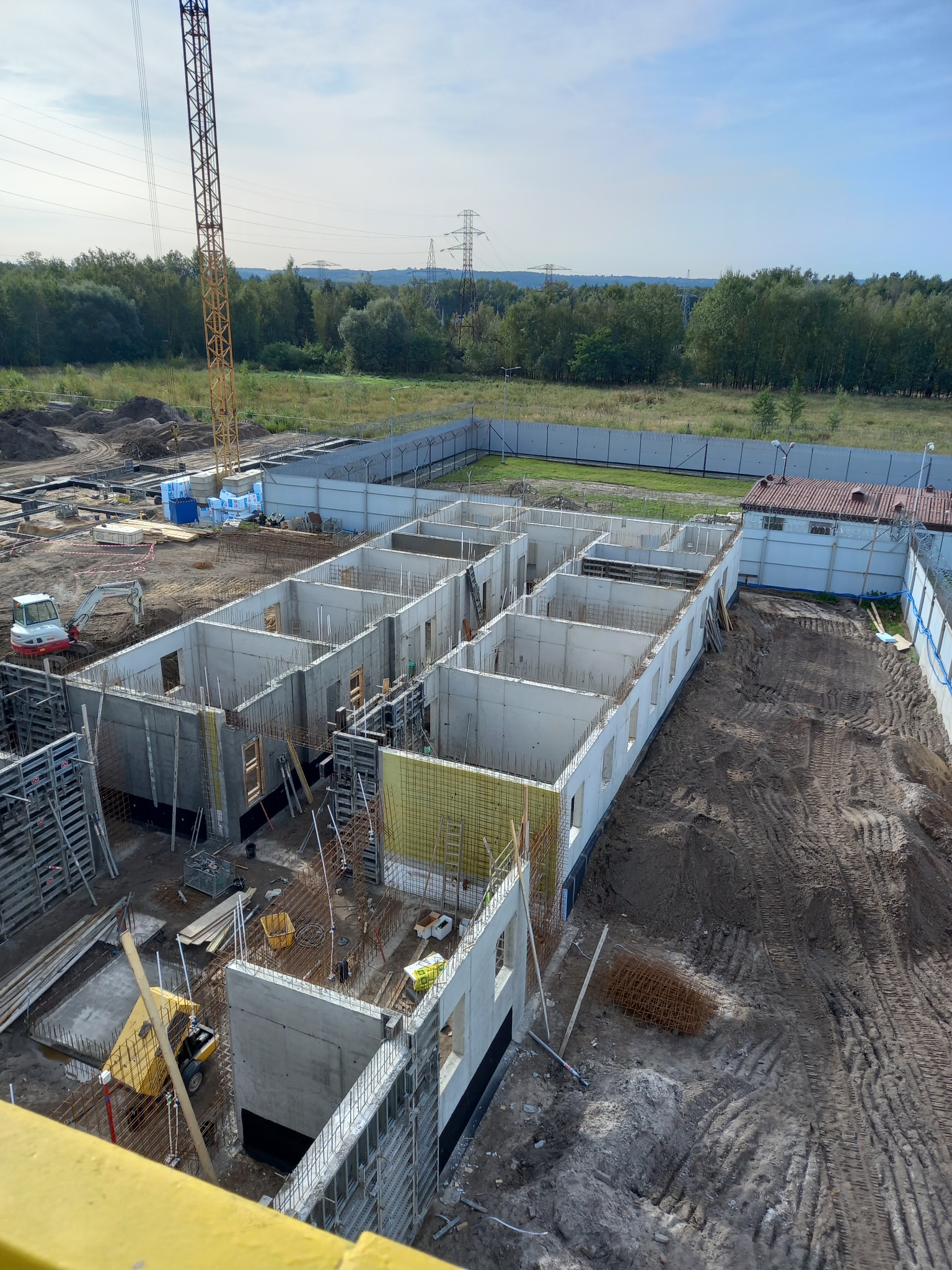 Budowa w toku: widok z góry na fundamenty segmentów w stanie surowym otwartym, zbrojenie, szalunki, mała koparka i żuraw na placu budowy. Otoczenie: drzewa, ogrodzenie.
