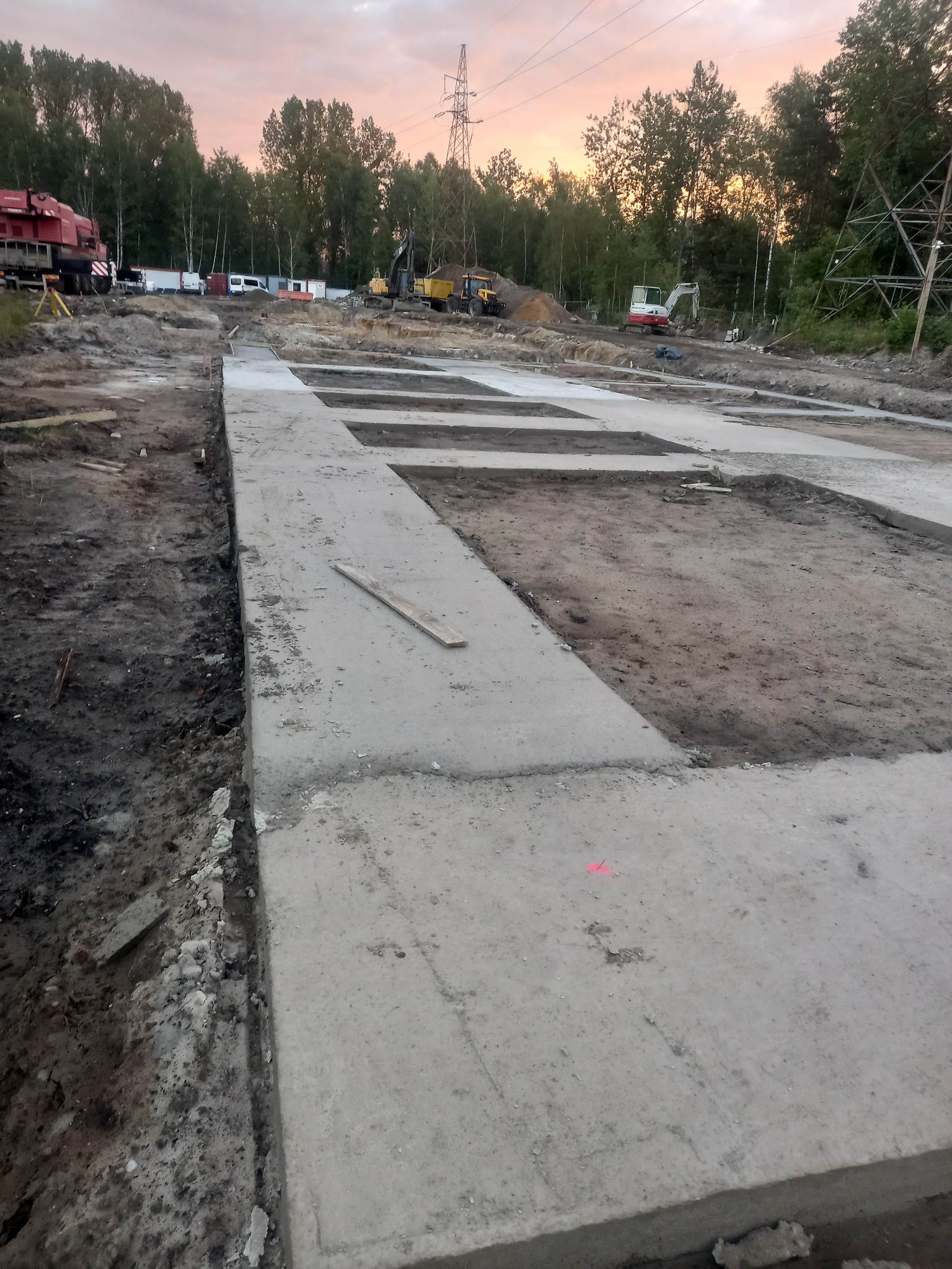 Zalane betonem fundamenty pod budynek, widoczne wykopy i ciężki sprzęt na tle drzew i zachodzącego słońca. W tle słupy wysokiego napięcia.