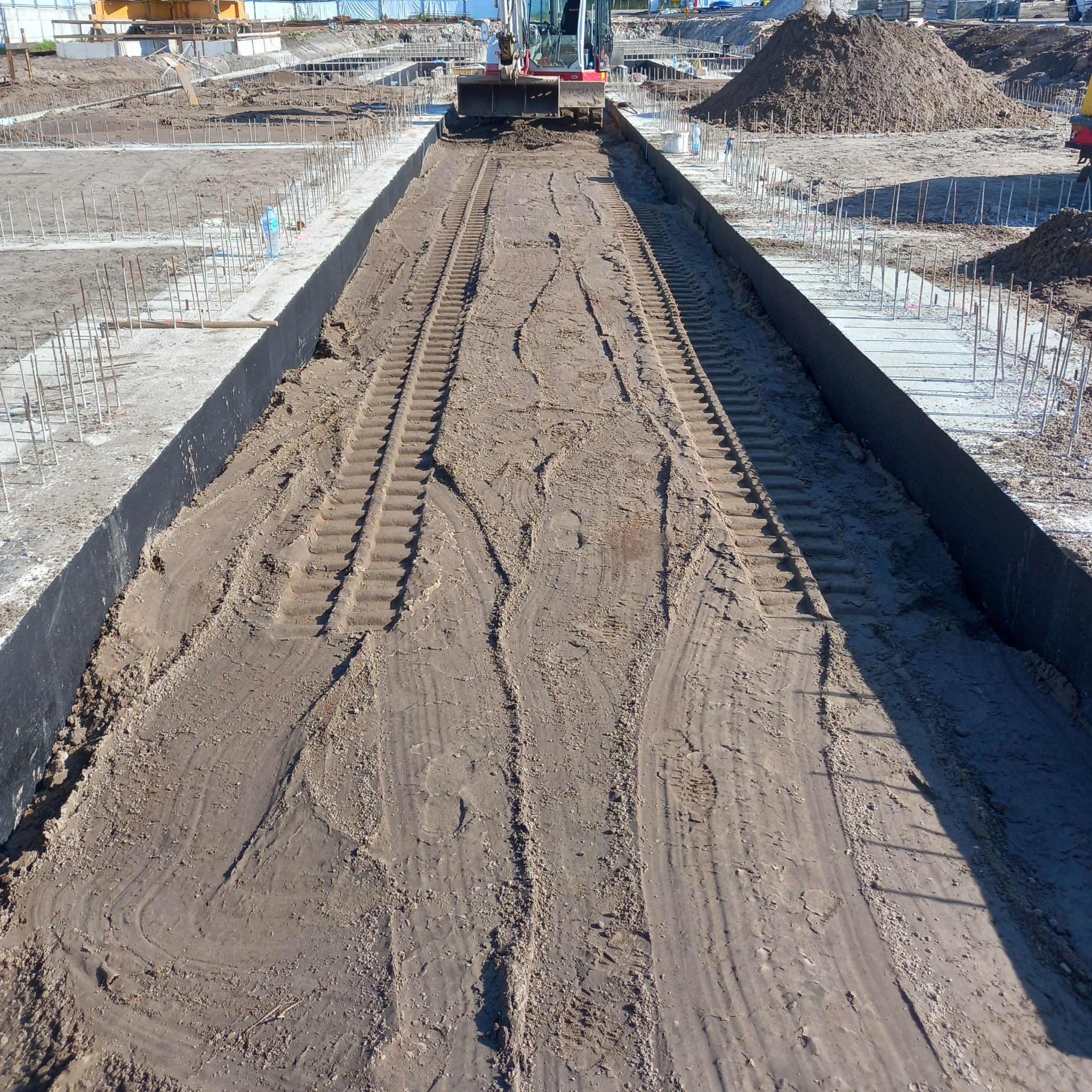 Wykop pod fundamenty z widocznymi śladami gąsienic koparki, betonowe ściany wykopu z wystającymi prętami zbrojeniowymi. W tle hałda ziemi i pracująca koparka.