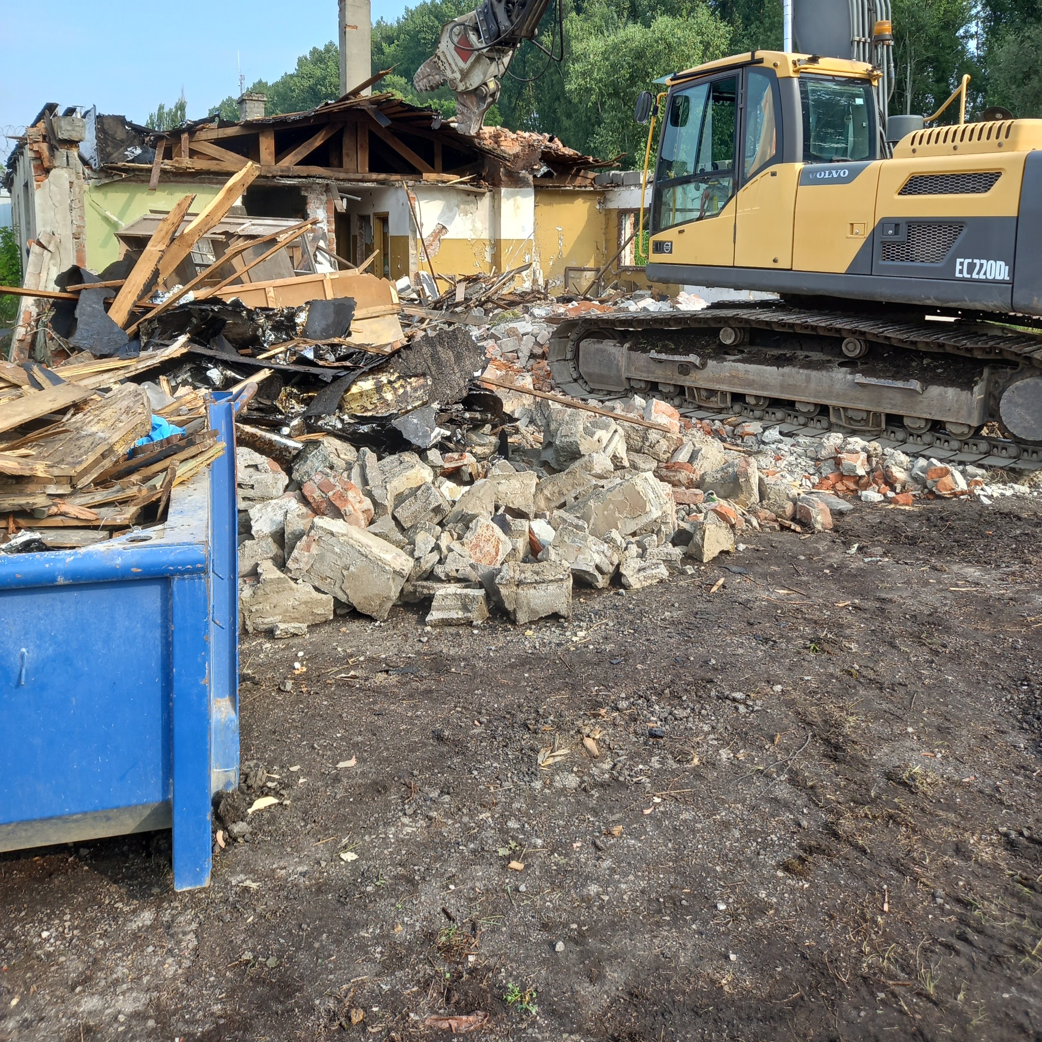 Żółta koparka Volvo EC220DL podczas rozbiórki budynku, z niebieskim kontenerem na gruz w tle. Widoczne porozrzucane cegły i elementy konstrukcyjne.