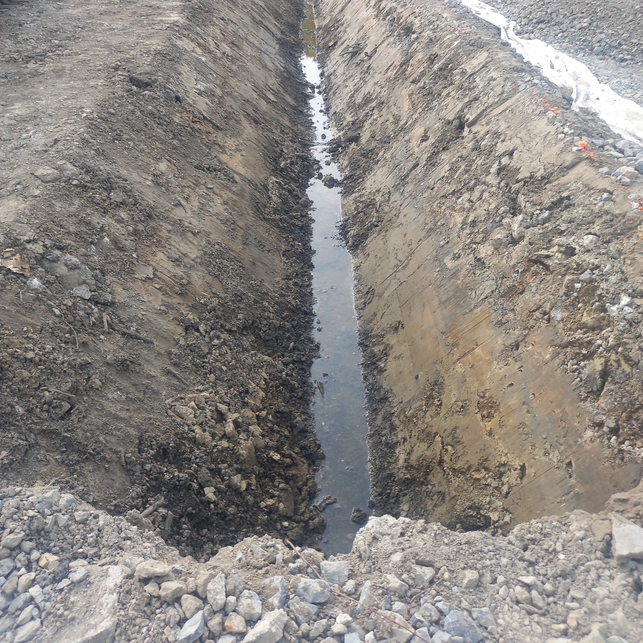 Długi, wąski wykop wypełniony wodą, otoczony ziemią i kamieniami. Widoczna biała tkanina na jednym z brzegów. Ujęcie z góry.