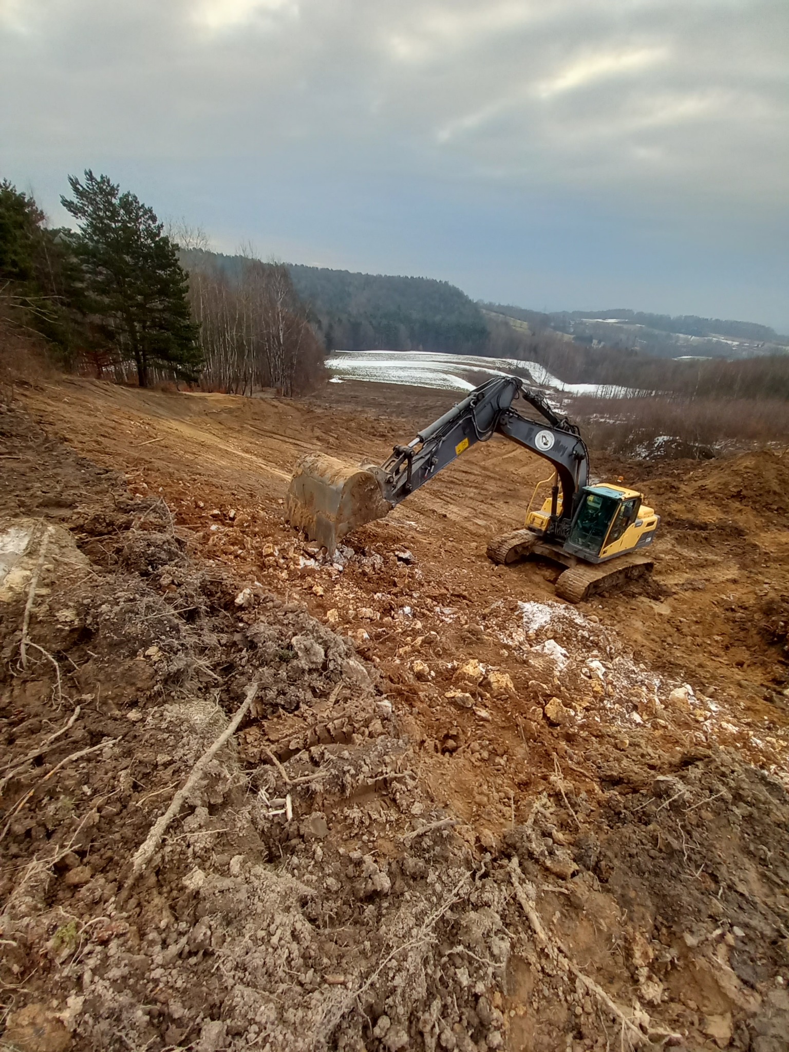Żółta koparka Volvo na zboczu wzgórza, w tle las i pole pokryte śniegiem pod pochmurnym niebem. Maszyna w trakcie prac ziemnych, widoczne ślady wykopów.