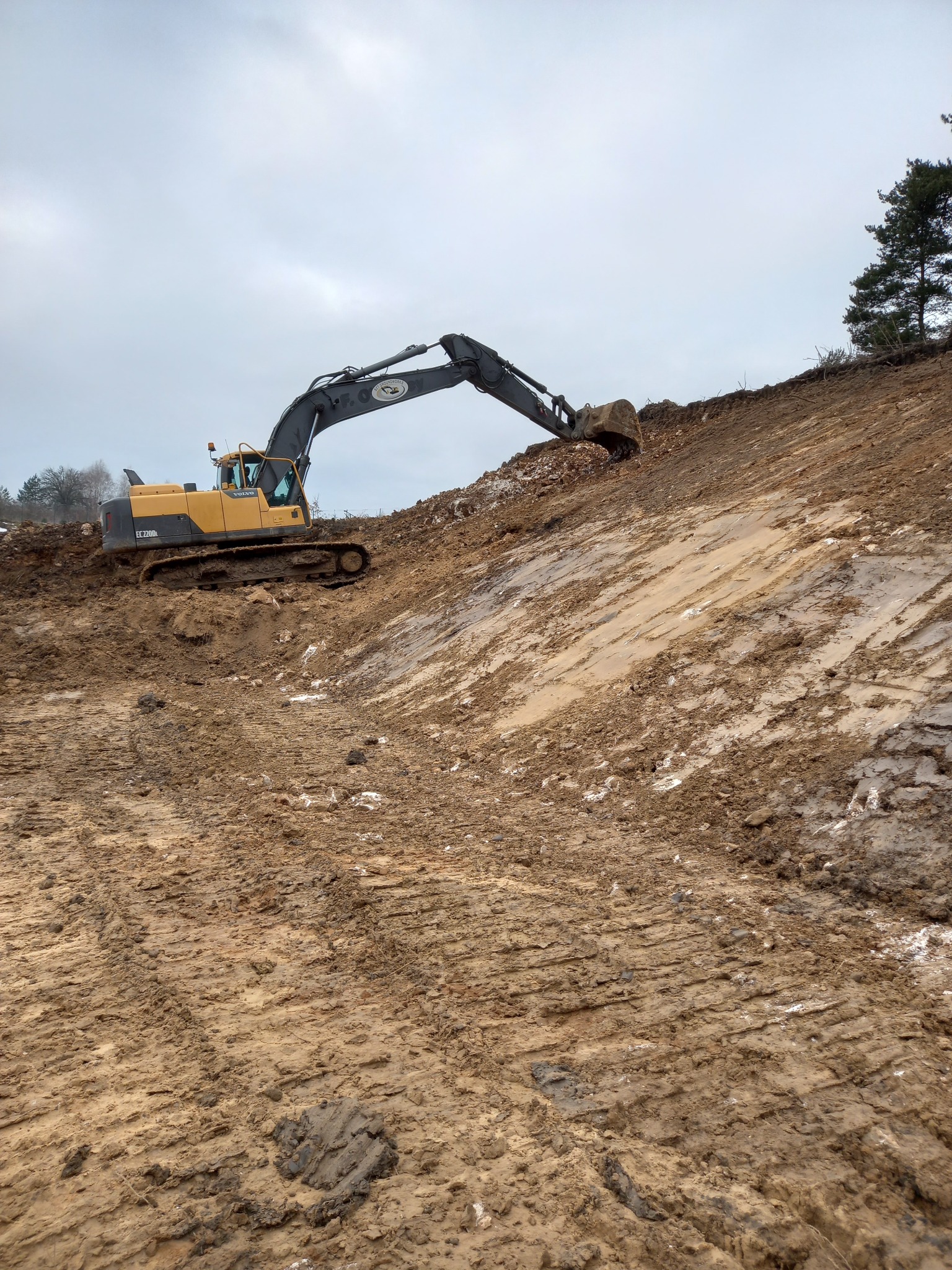 Żółta koparka Volvo EC220DL na zboczu ziemi, ślady opon w błocie, pochmurne niebo, drzewo na szczycie wzgórza. Prace ziemne w toku.