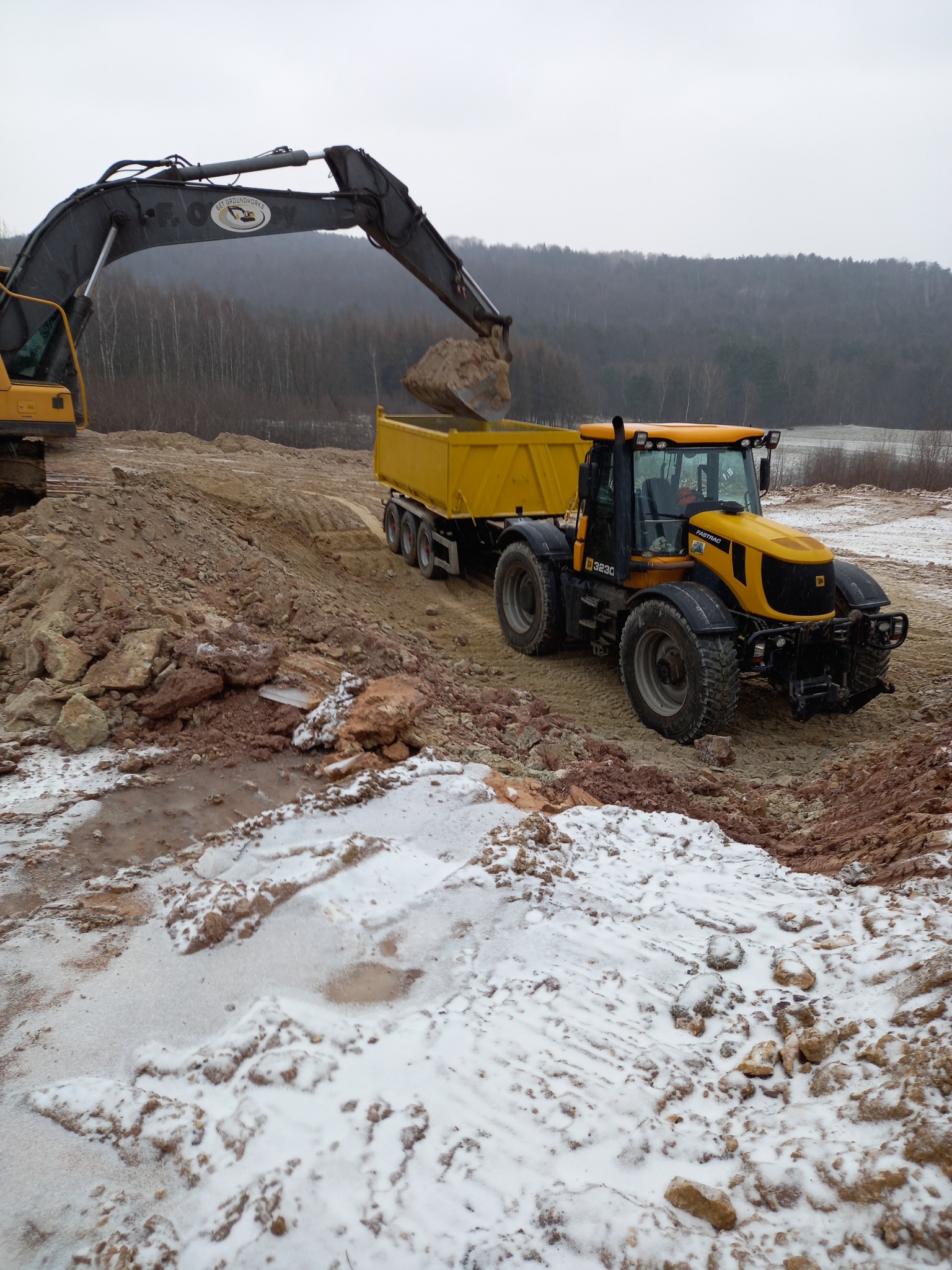 Żółty traktor JCB 3230 z przyczepą, ładowarka Volvo z łyżką nabiera ziemię. Prace ziemne w terenie z resztkami śniegu i błota, zalesione wzgórze w tle.