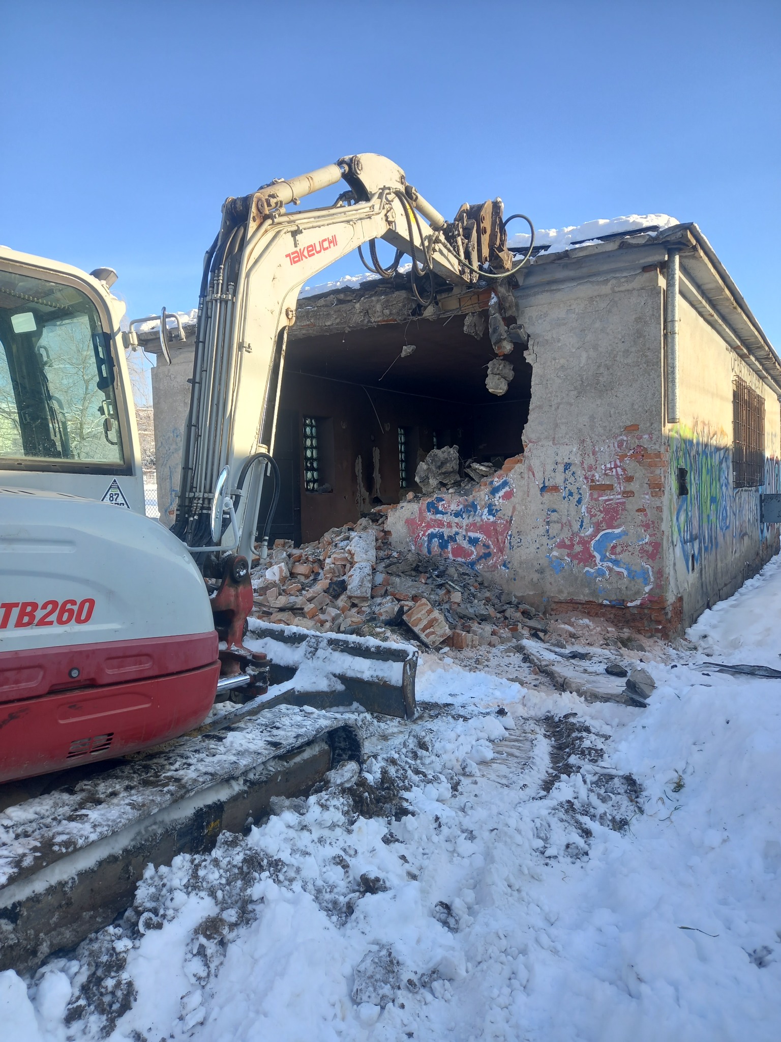 Rozbiórka budynku z graffiti w zimowej scenerii. Koparka Takeuchi TB260 w trakcie pracy, gruz leży na śniegu. Widoczne zniszczone ściany i fragment dachu.