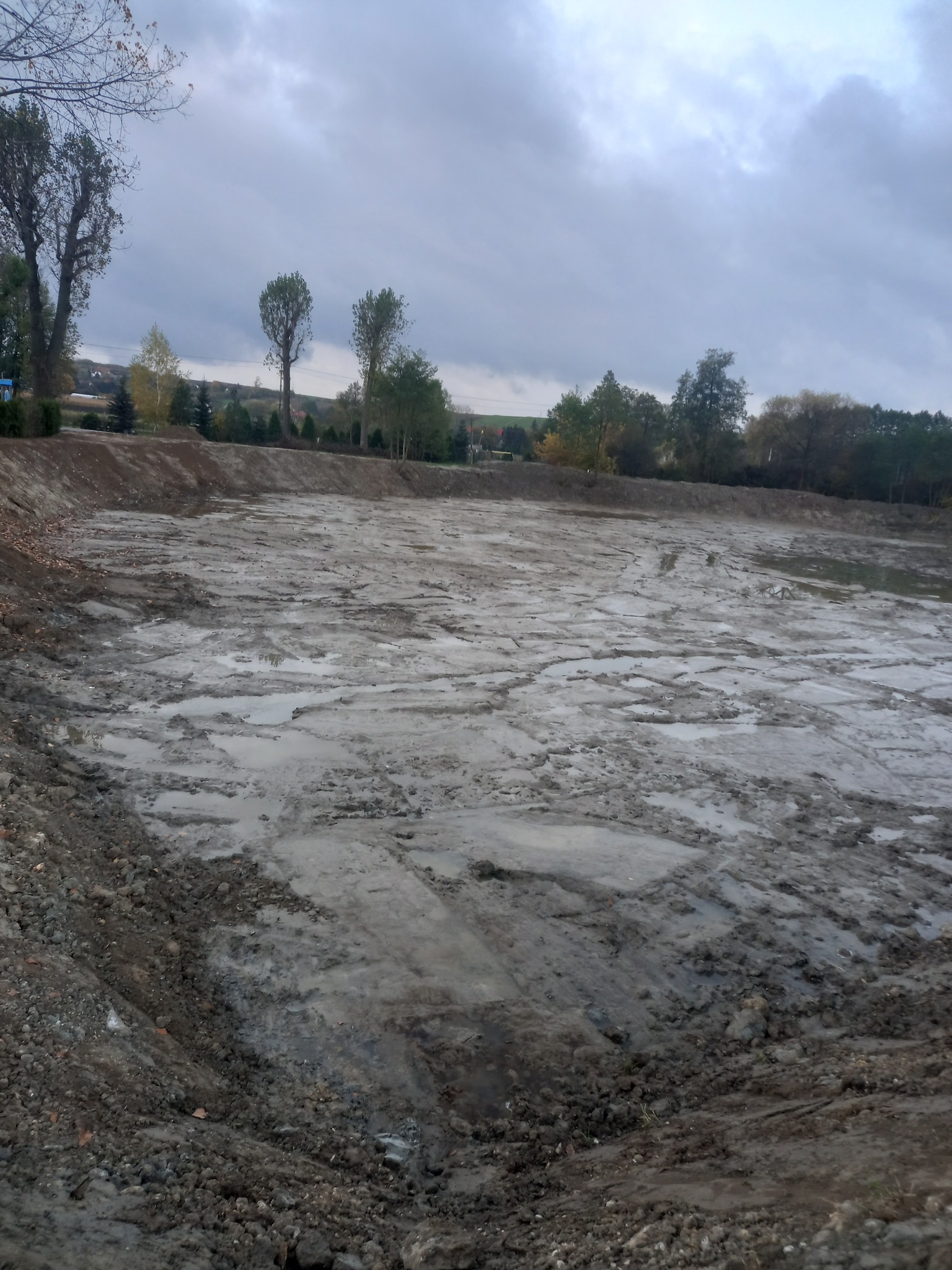 Wykopany, błotnisty teren pod budowę stawu w pochmurny dzień. Widoczne ślady maszyn i nierówności terenu, drzewa w tle.