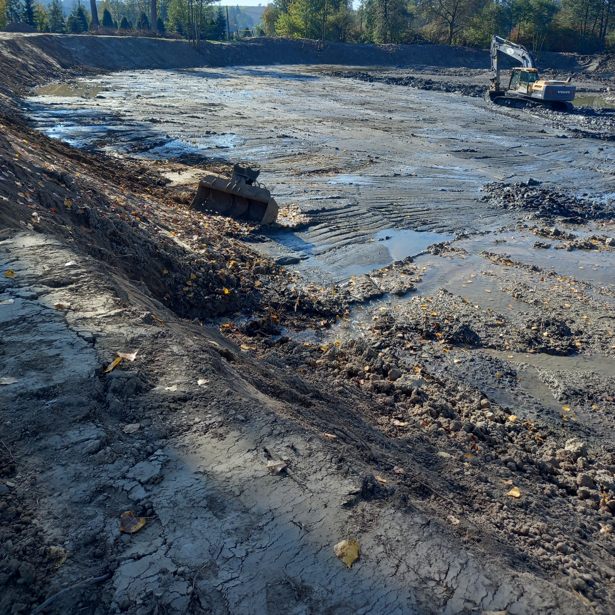 Wykopany staw w trakcie prac ziemnych, z widocznym błotem, wodą i koparką VOLVO w tle. Brzeg wykopu nierówny, z liśćmi.
