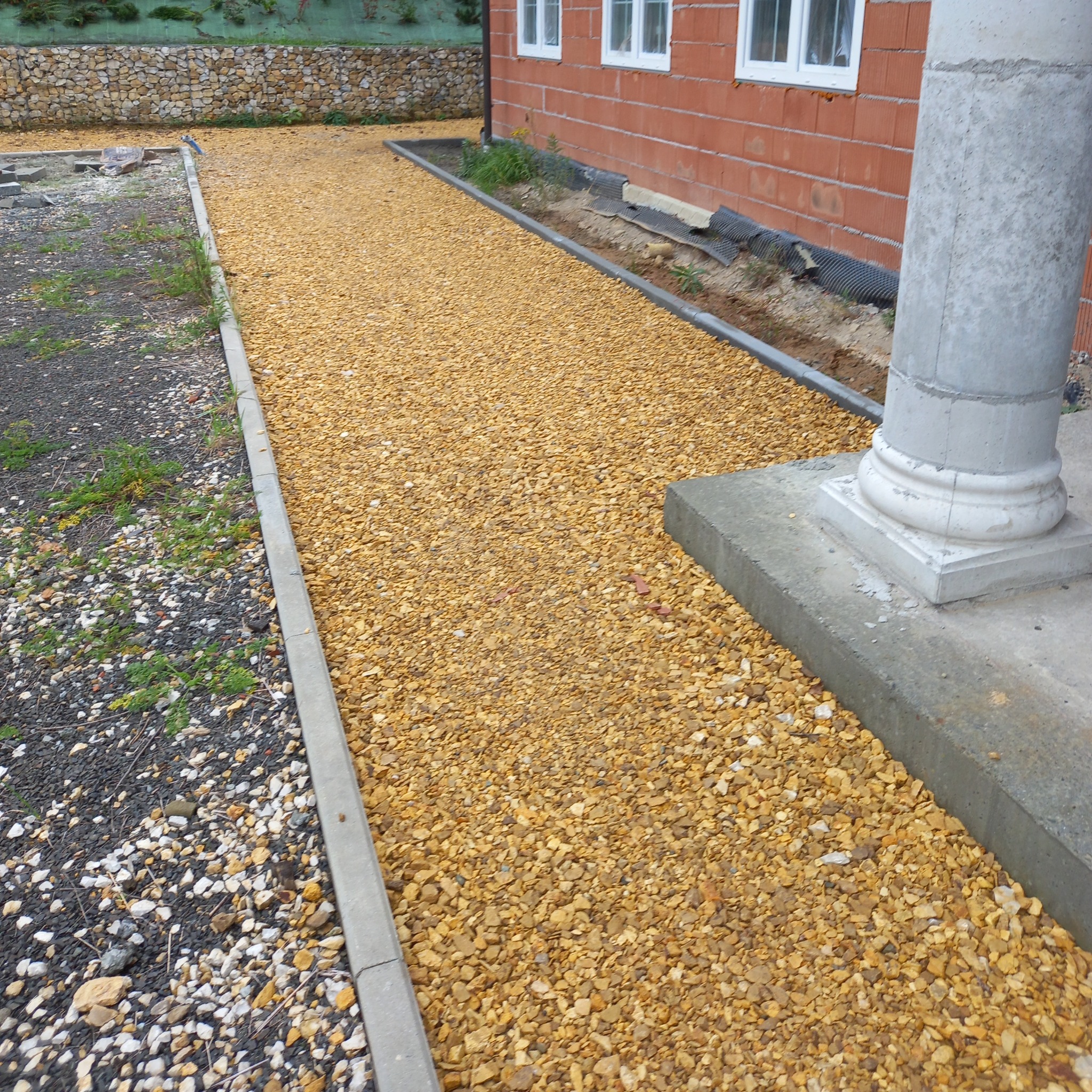 Utwardzona, złocista ścieżka z kamienia ozdobnego obok budynku z cegły, z widocznym betonowym obrzeżem i fragmentem kolumny. Część ścieżki z ciemniejszym kamieniem.