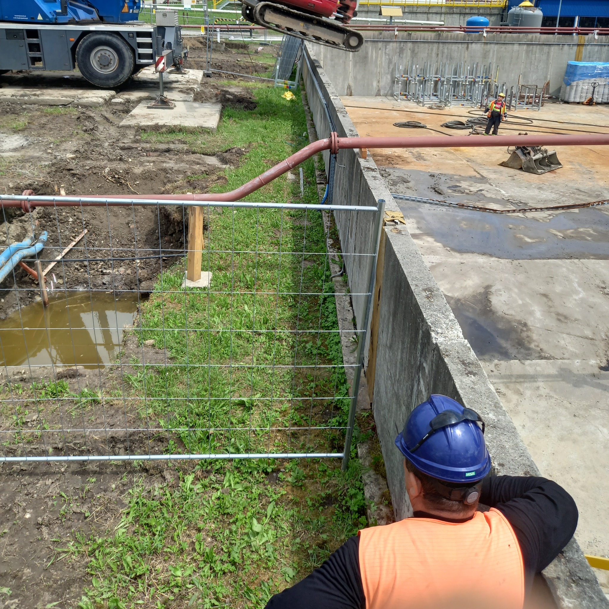 Instalacja rur na placu budowy z wykopem, widoczny dźwig i pracownik w kasku. Częściowo ogrodzony teren. Prace ziemne w toku, z widocznymi elementami infrastruktury.