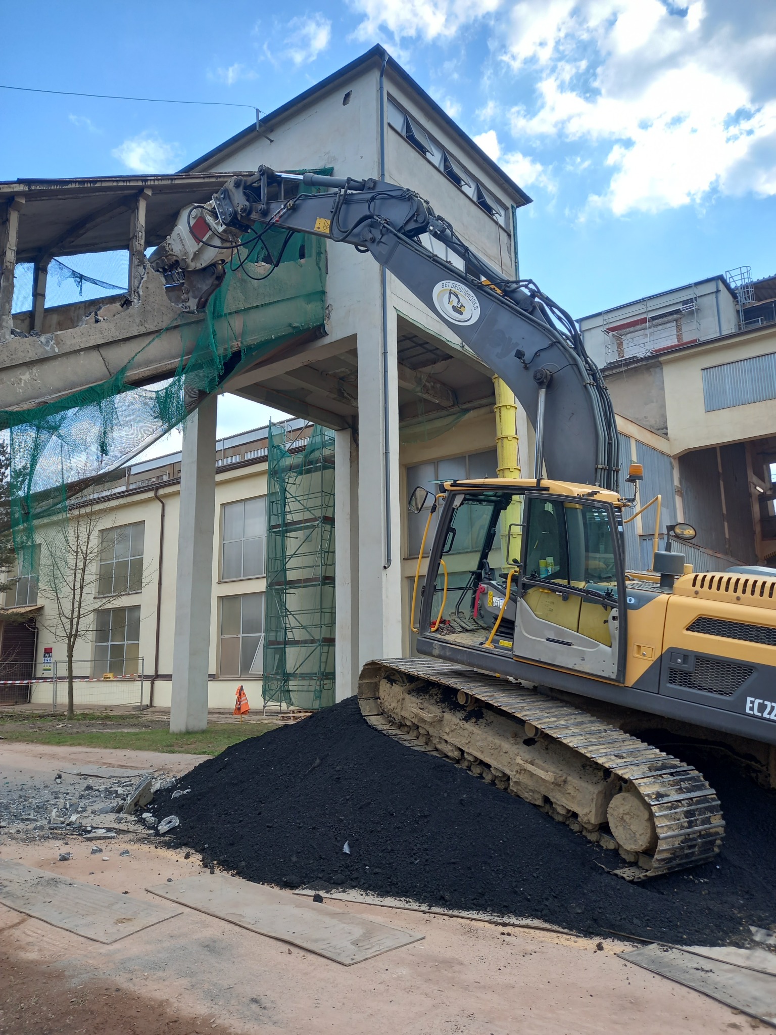 Żółty koparko-ładowarka z hydraulicznymi nożycami tnie betonowy element konstrukcyjny budynku w trakcie rozbiórki. Widoczne zabezpieczenia siatką.