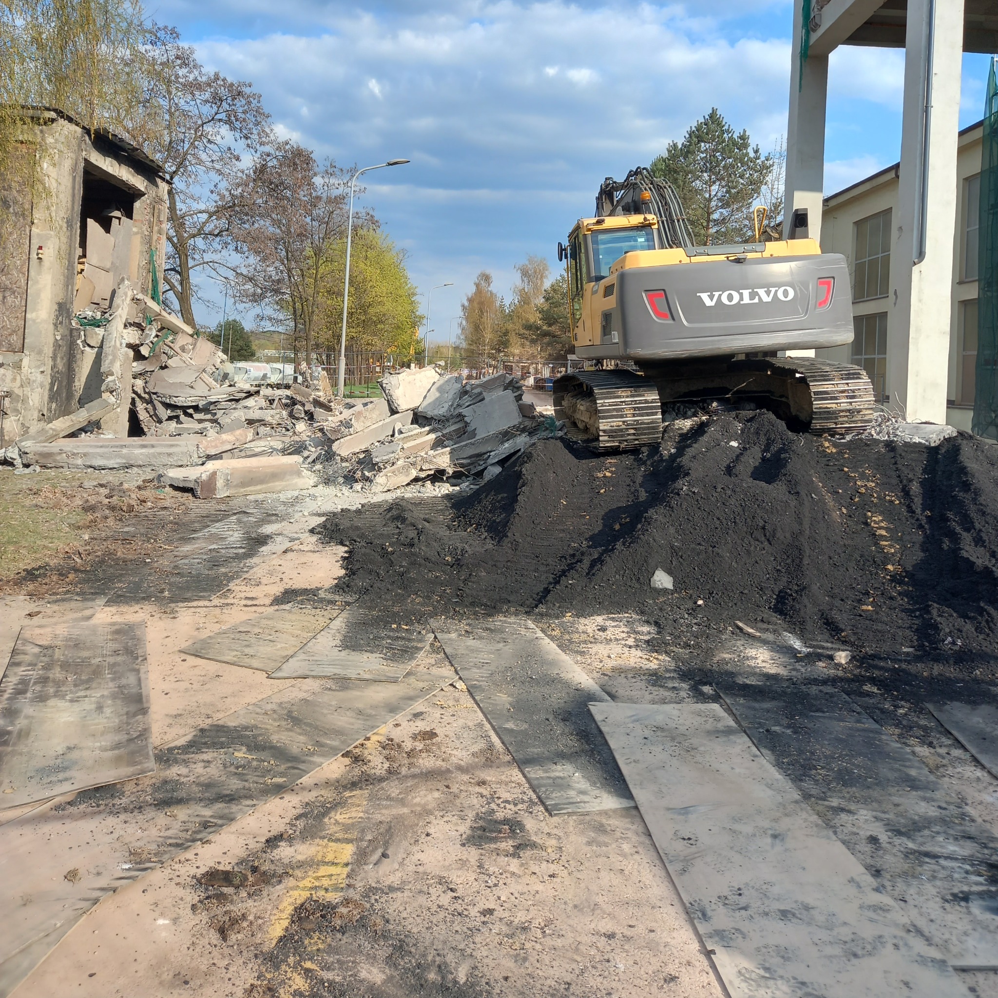 Żółta koparka Volvo na hałdzie ziemi, w tle gruzowisko budynku po rozbiórce. Wokół płyty drogowe, drzewa i budynki. Błękitne niebo z chmurami.