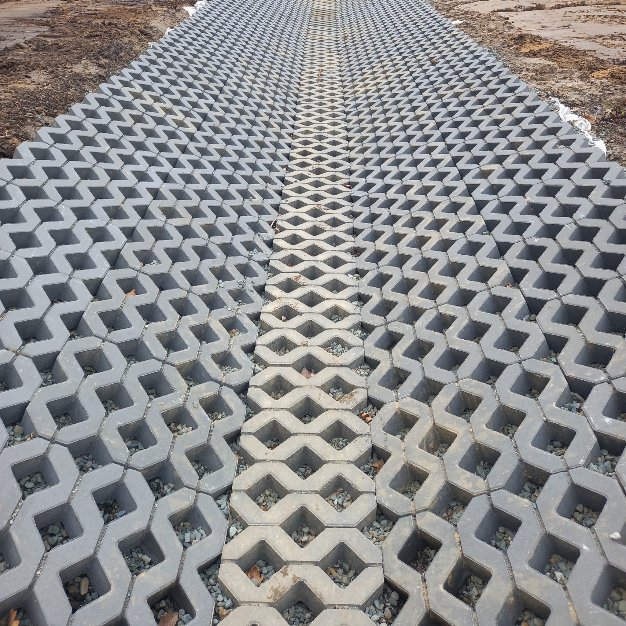 Ułożona szara ażurowa nawierzchnia z wypełnieniem żwirowym na gruncie, tworząca ścieżkę. Widok z góry, ukazujący regularny wzór i strukturę materiału.