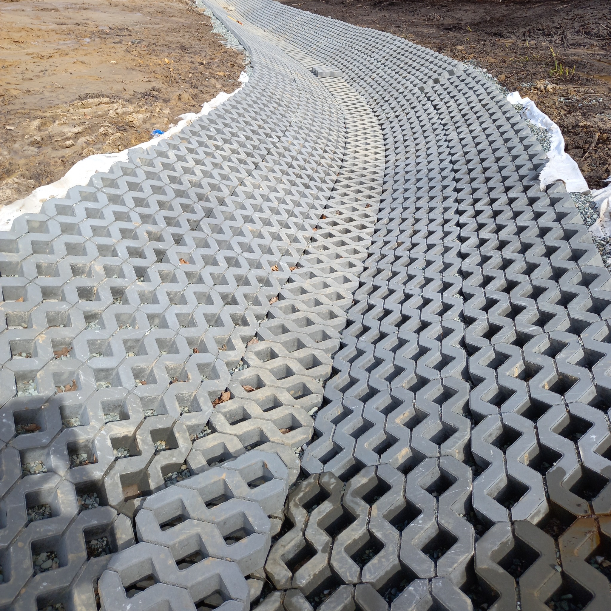 Ułożona, kręta ścieżka z szarych, ażurowych płyt betonowych na podsypce żwirowej. Widoczna ziemia po bokach. Elementy ułożone w zygzakowaty wzór.