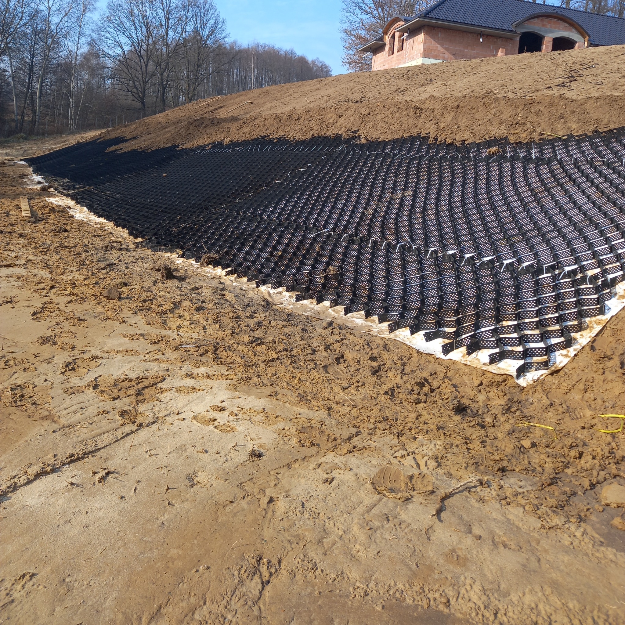 Geokrata na zboczu w trakcie prac ziemnych. W tle budynek w stanie surowym i drzewa. Ujęcie z dołu, podkreślające strukturę geokraty.