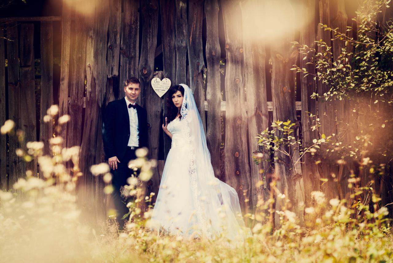 Para młoda, pan młody w czarnym smokingu i panna młoda w białej sukni ślubnej z welonem, pozują na tle starej, drewnianej stodoły z sercem wiszącym na drzwiach, rozmyte kwiaty na pierwszym planie.