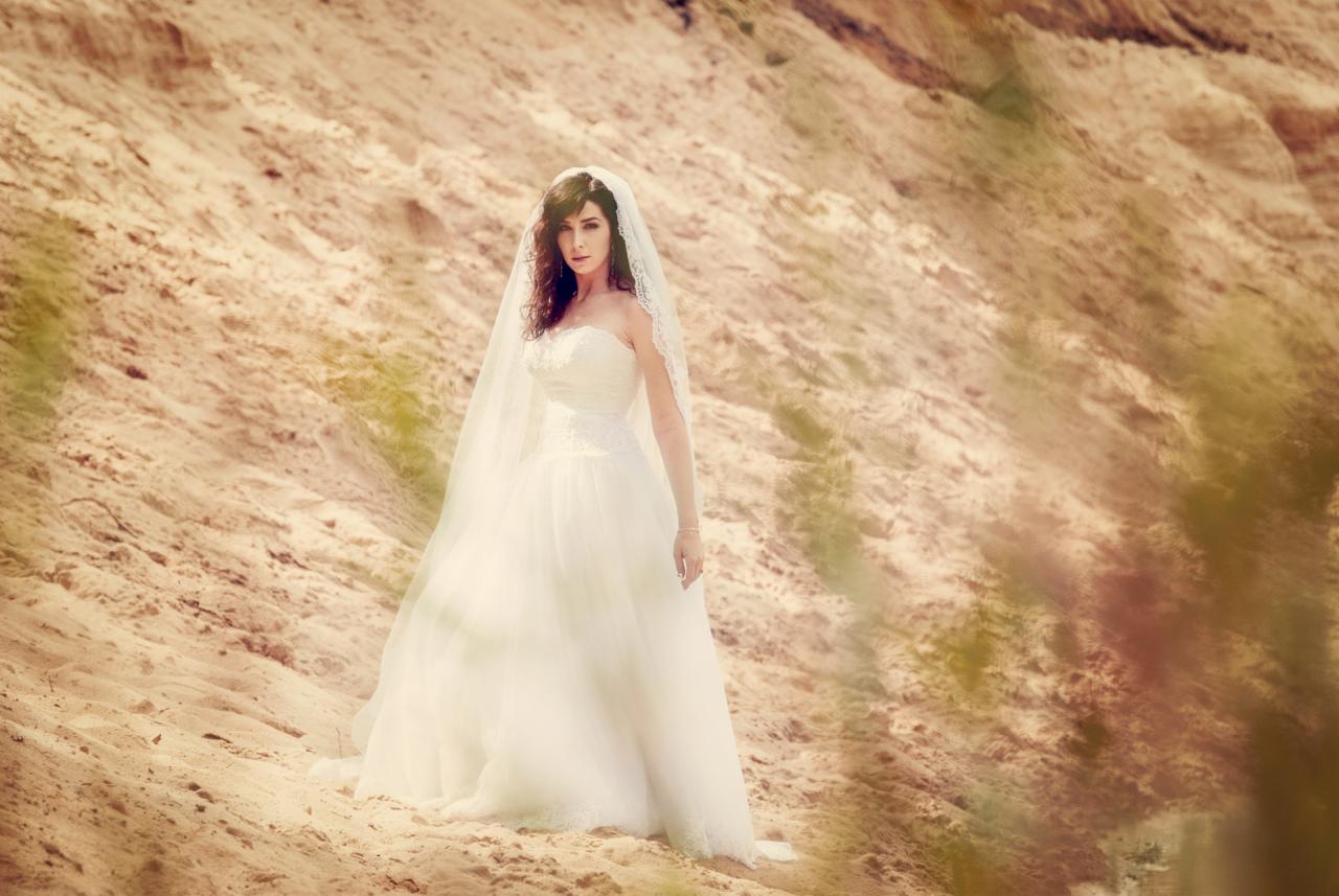 Panna młoda w sukni ślubnej i welonie pozuje na tle piaszczystej wydmy w słoneczny dzień, z delikatnym efektem vintage.