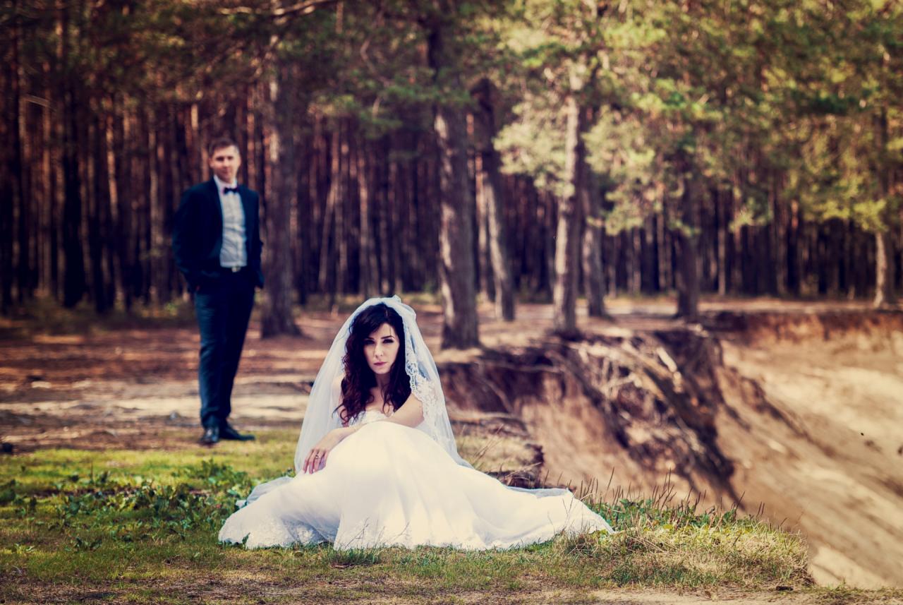Panna młoda w białej sukni i welonie siedzi na trawie na skraju lasu, w tle widoczny pan młody w garniturze.