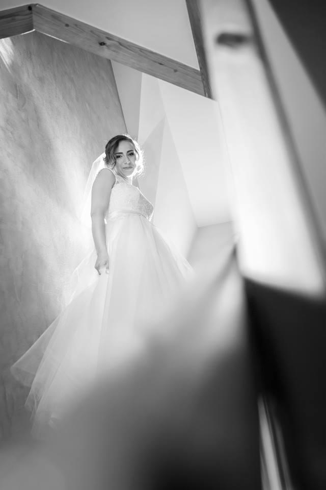 Czarno-białe zdjęcie panny młodej w sukni ślubnej z welonem, stojącej na schodach w nowoczesnym wnętrzu, oświetlonej naturalnym światłem wpadającym przez okno.