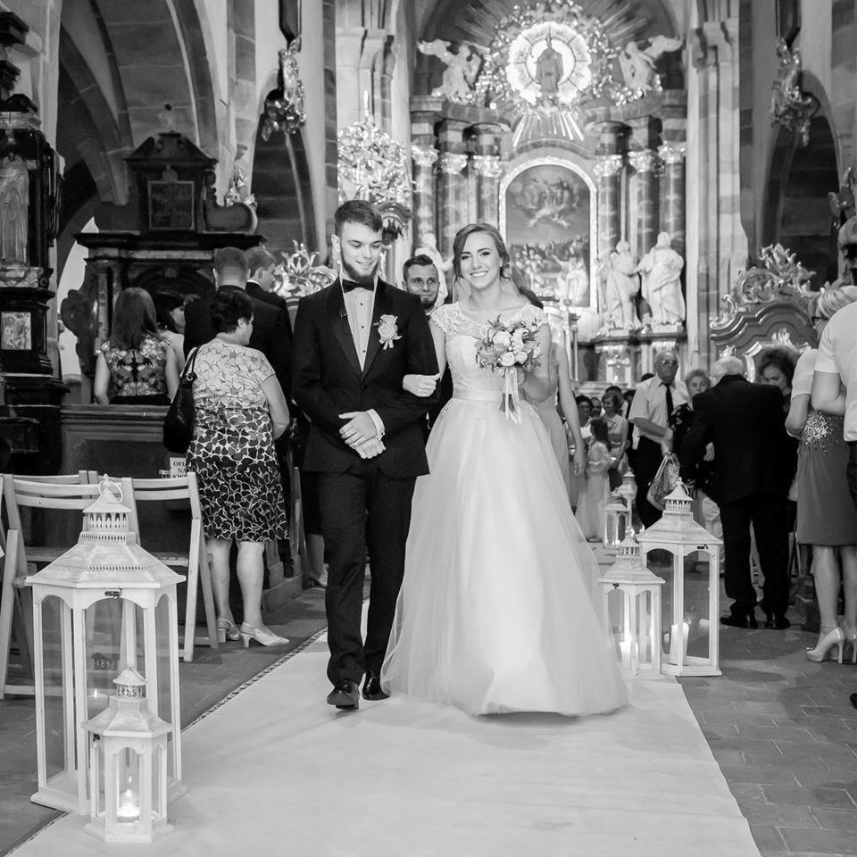 Czarno-białe zdjęcie nowożeńców idących w kościele, panna młoda w sukni z bukietem, pan młody w smokingu, w tle ołtarz i goście, w pierwszym planie lampiony.