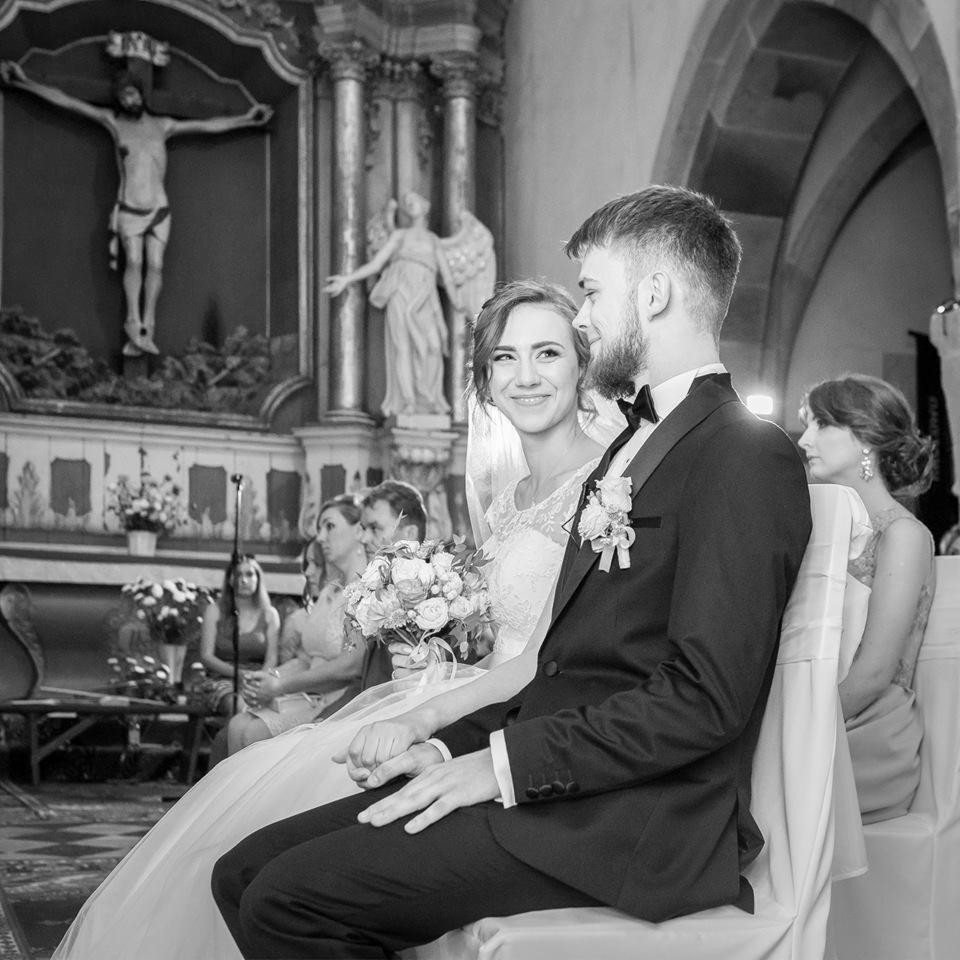 Czarno-białe zdjęcie nowożeńców siedzących w kościele podczas ceremonii ślubnej, z widocznym krucyfiksem i posągiem anioła w tle.