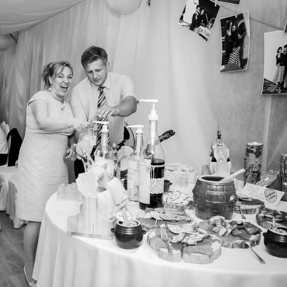 Czarno-białe zdjęcie pary nalewającej napój z dzbanka na stole z przekąskami, drewnianymi dekoracjami i zdjęciami w tle.