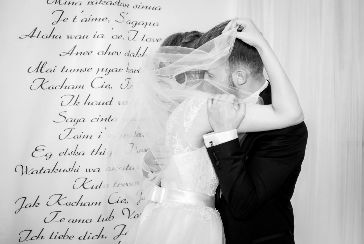 Czarno-białe zdjęcie pary młodej w objęciach, panna młoda w koronkowej sukni i welonie, pan młody w czarnym garniturze, tło stanowi ściana z napisem 'Kocham Cię' w różnych językach.