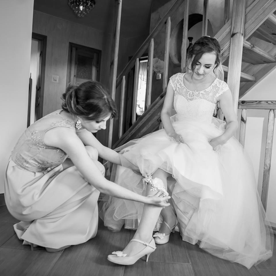 Czarno-białe zdjęcie panny młodej siedzącej na schodach, druhna zakłada podwiązkę na jej nodze, widoczne buty ślubne i fragment sukni z koronkową górą.