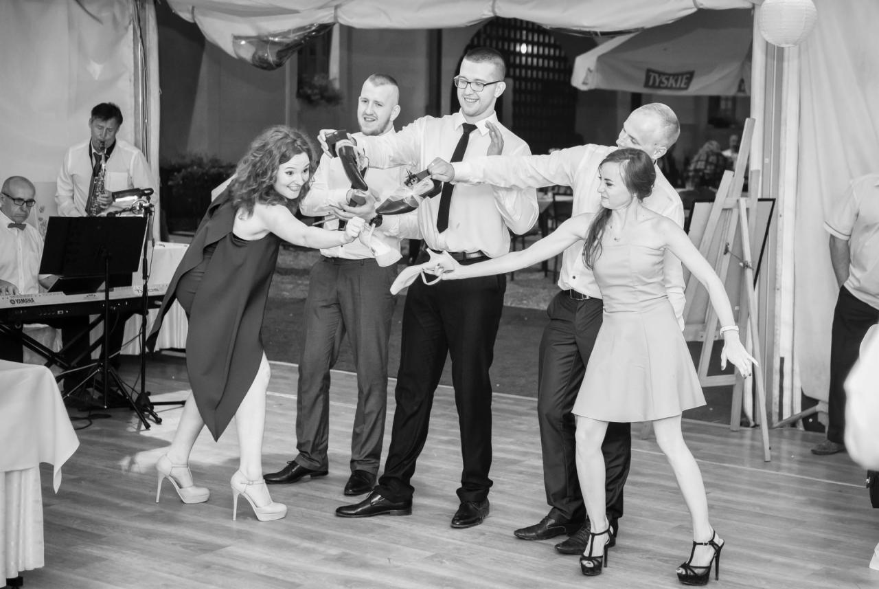 Czarno-białe zdjęcie z oczepin weselnych: panna młoda i jedna z druhen próbują odebrać buty trzymane przez czterech mężczyzn, w tle zespół muzyczny i parasol Tyskie.