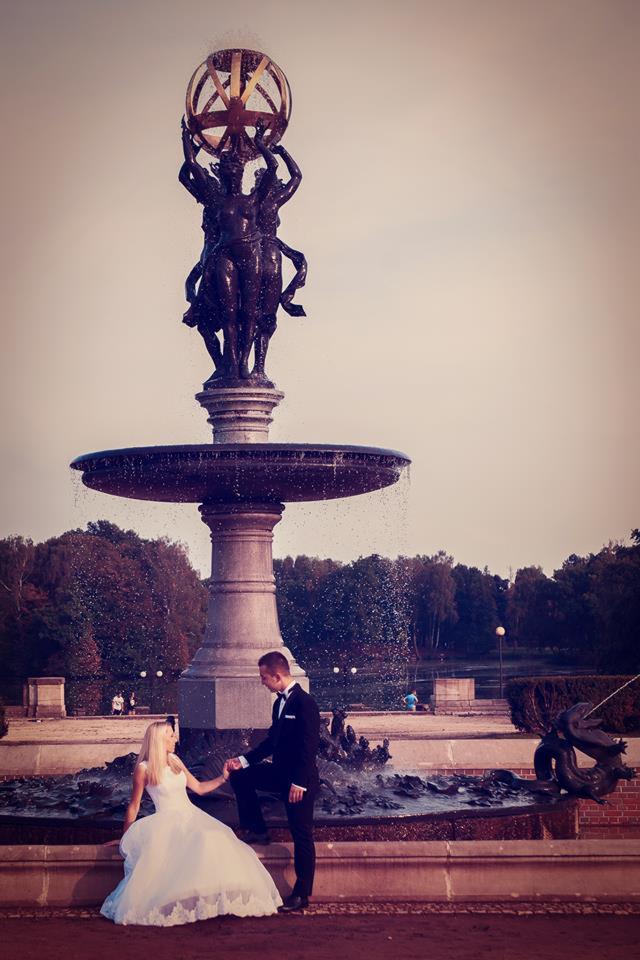 Para młoda w strojach ślubnych pozuje przy fontannie z rzeźbą w parku miejskim w słoneczny dzień.
