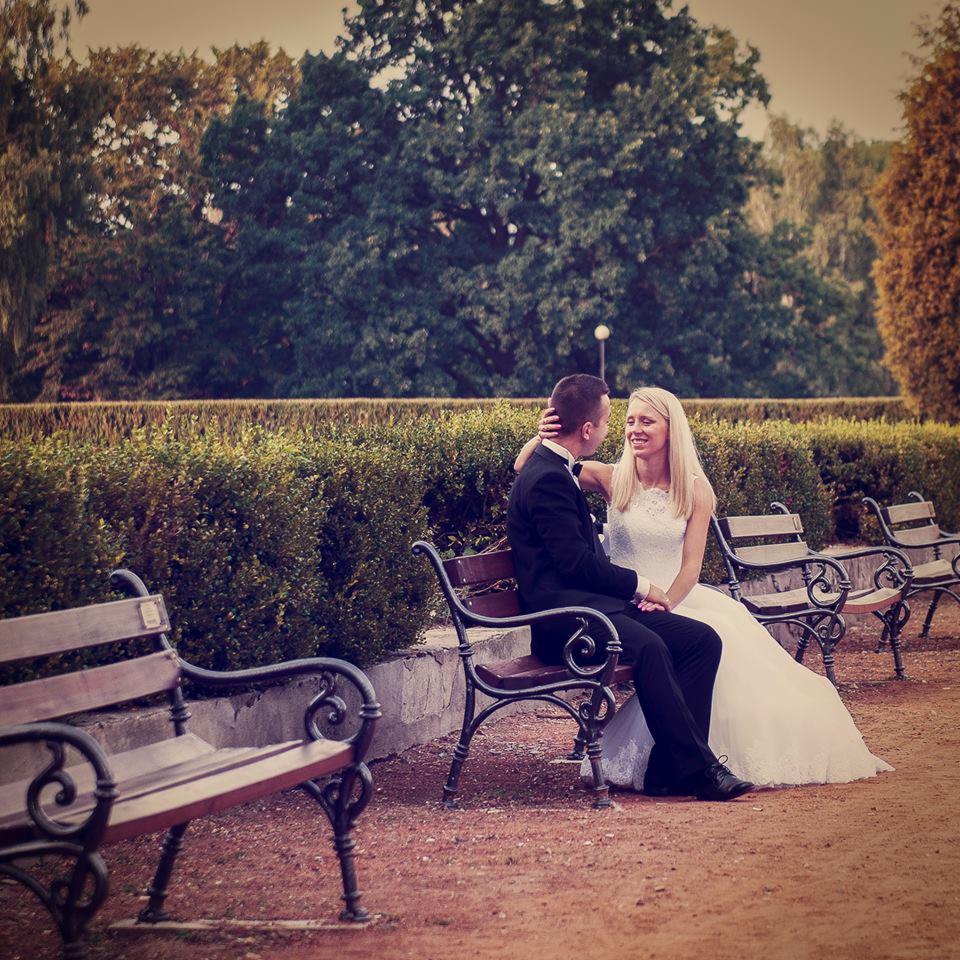 Para młoda siedząca na ławce w parku, panna młoda w białej sukni z koronkową górą, pan młody w czarnym smokingu z muszką, uśmiechnięci i patrzący na siebie.