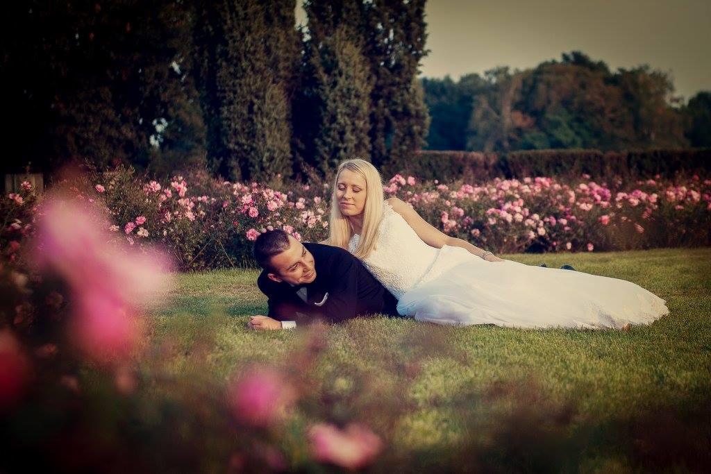Para młoda leżąca na trawie w ogrodzie różanym, panna młoda w białej sukni, pan młody w czarnym smokingu, romantyczna sesja plenerowa.