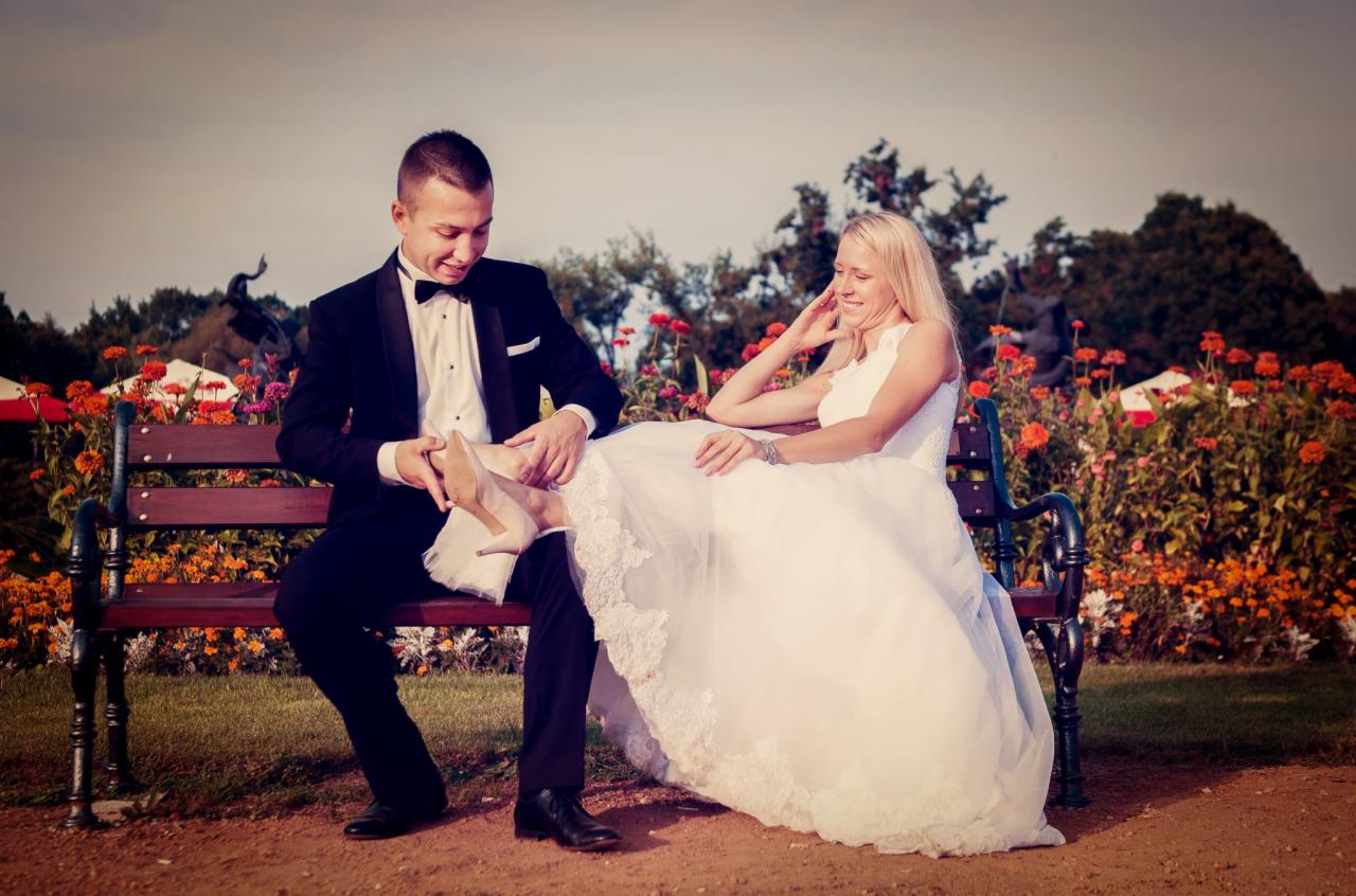 Pan młody w smokingu zakłada pantofel na stopę uśmiechniętej panny młodej w białej sukni, siedzącej na ławce w parku z pomarańczowymi kwiatami w tle.