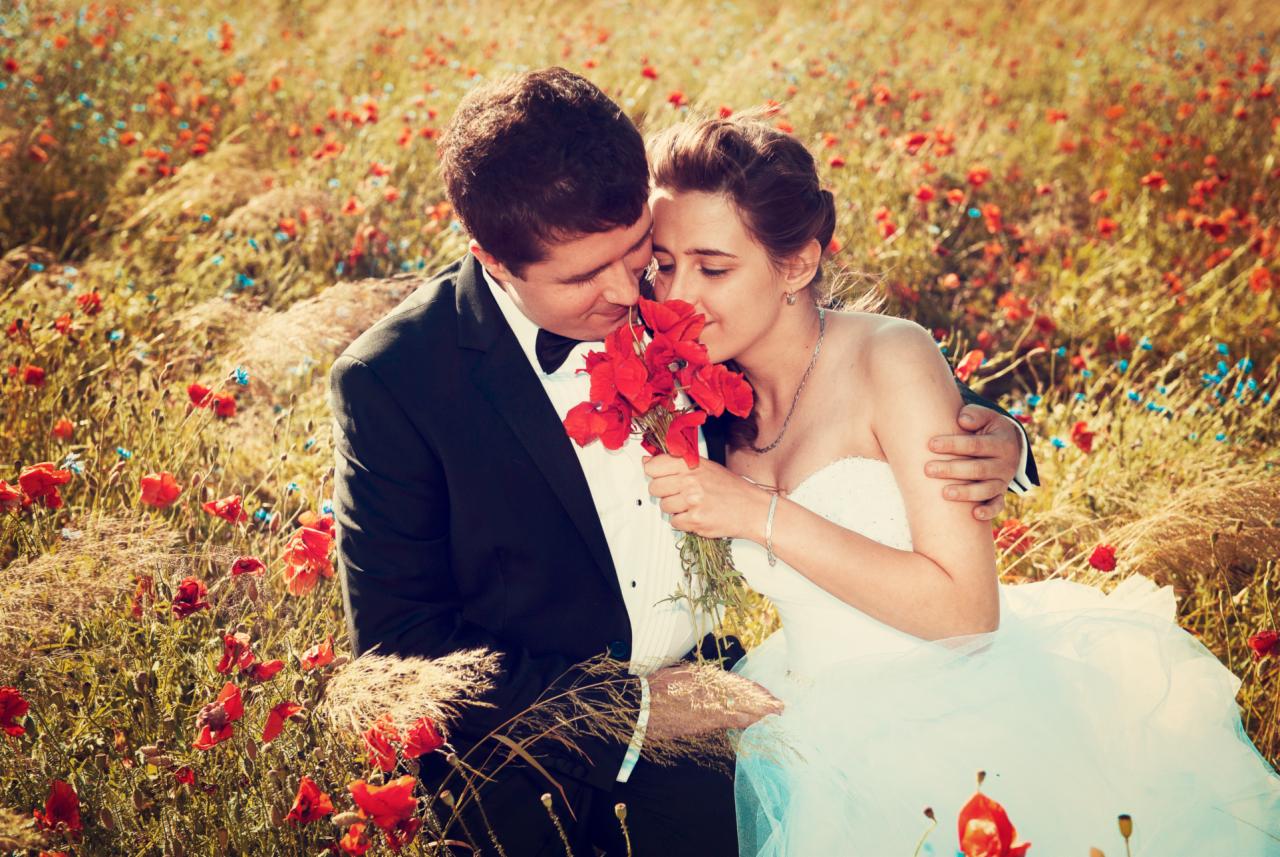 Para młoda w polu maków, panna młoda wącha bukiet czerwonych kwiatów, pan młody obejmuje ją ramieniem.