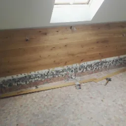 Montaż drewnianej boazerii na ścianie poddasza z widocznymi przewodami elektrycznymi, mozaikowa podłoga w trakcie remontu, narzędzia pracy i listwy przypodłogowe.