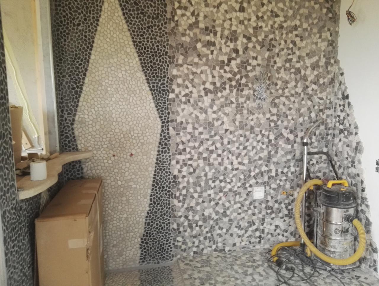 Mozaika z kamyczków w odcieniach szarości i bieli, tworząca wzór na ścianie i podłodze łazienki, z widocznym odkurzaczem przemysłowym i kartonem.