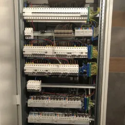 Otwarta szafa elektryczna z licznymi bezpiecznikami, przekaźnikami i przewodami ułożonymi wewnątrz, z widocznymi złączkami w kolorach niebieskim, zielonym i żółtym.