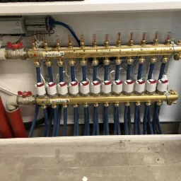 Rozdzielacz ogrzewania podłogowego Solter w szafce, z opisami stref (kuchnia, salon, sypialnia), widoczne zawory i niebieskie rurki, instalacja w trakcie montażu.