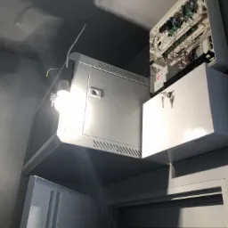 Otwarta szafa sterownicza z widocznymi elementami elektroniki, obok obudowa z kluczykami, oświetlone roboczą żarówką.