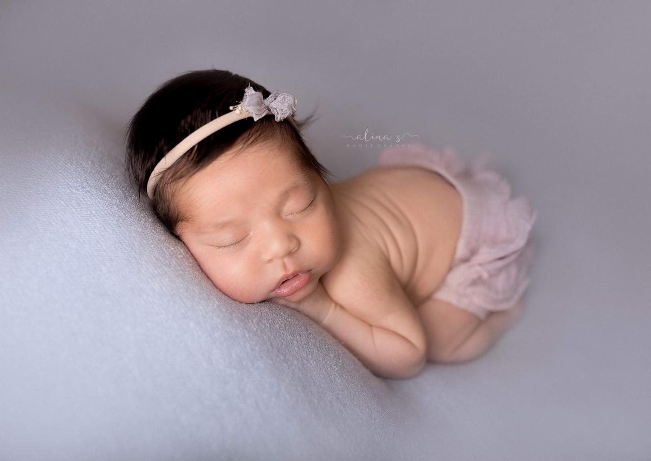 Śpiąca noworodka z ciemnymi włosami, ubrana w różowe falbanki i opaskę z kokardką, leżąca na błękitnym tle.