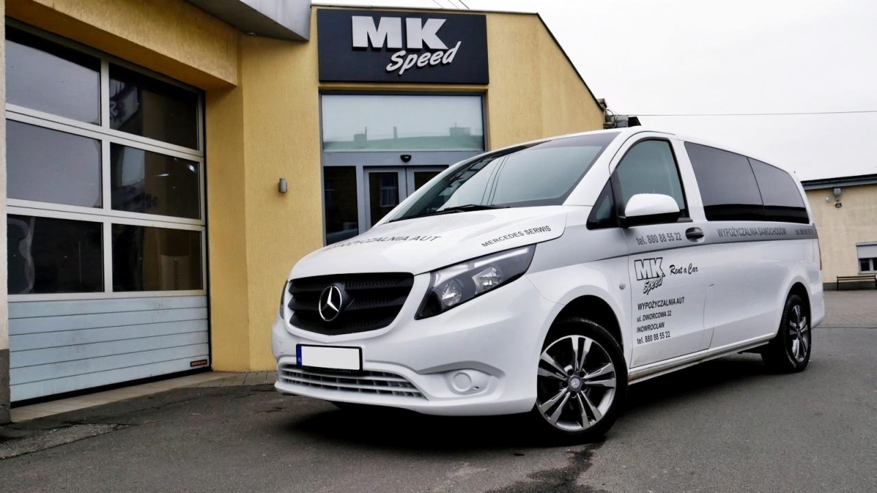 Biały Mercedes-Benz Vito z logo wypożyczalni samochodów MK Speed parkuje przed budynkiem firmy z żółtą elewacją i szarą bramą garażową.