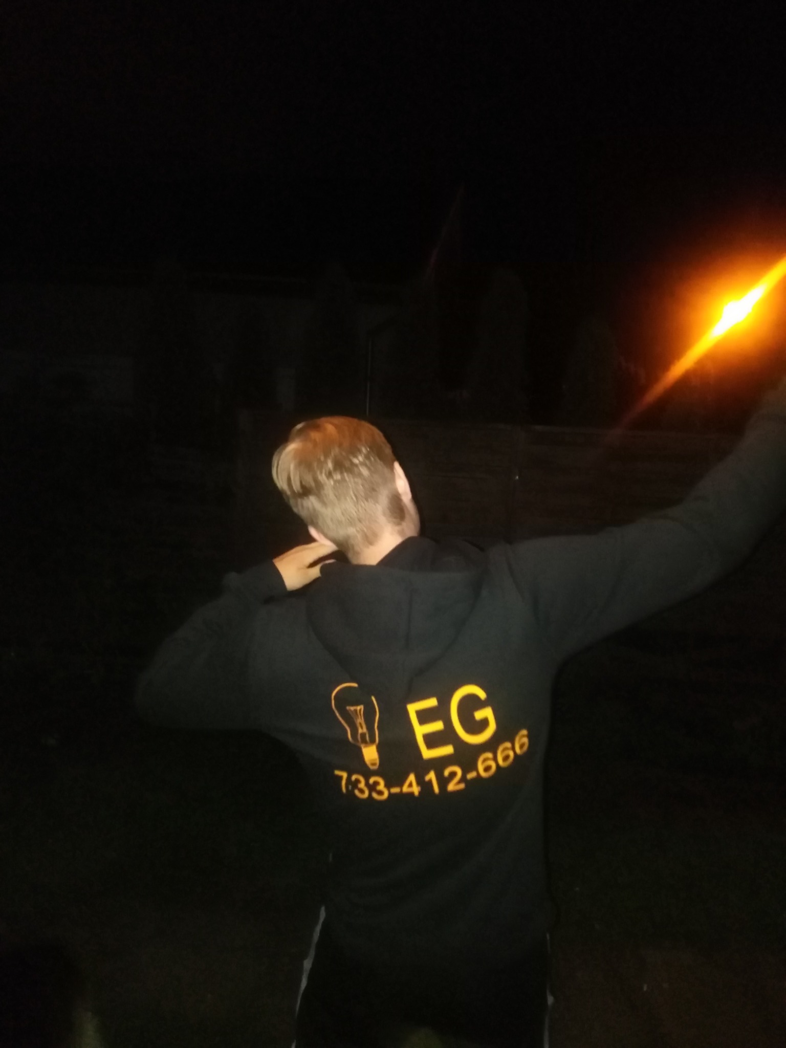 Mężczyzna w bluzie z kapturem z logo firmy z Głogowa, z motywem żarówki i numerem telefonu, pozuje w ciemności z wyciągniętymi rękami.