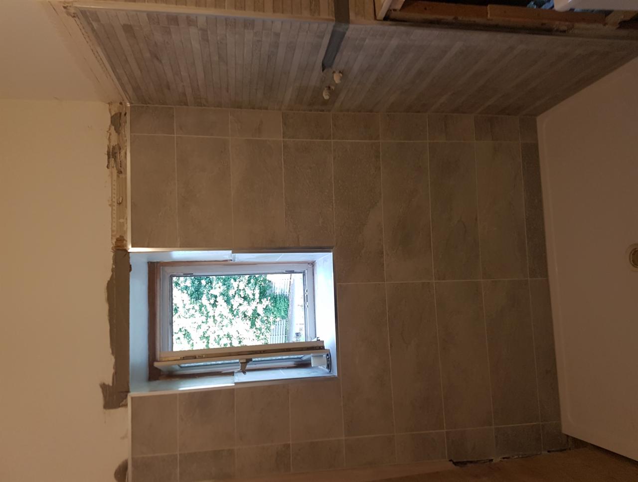Ściana i sufit w trakcie układania płytek ceramicznych w łazience, widoczne okno z widokiem na zieleń, niedokończone fugowanie przy oknie.