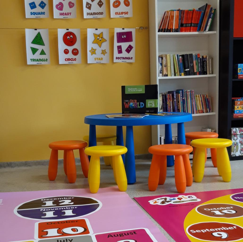 Sala lekcyjna z niebieskim stolikiem, kolorowymi stołeczkami, plakatami z kształtami i kalendarzem edukacyjnym na podłodze, książki na półce w tle.