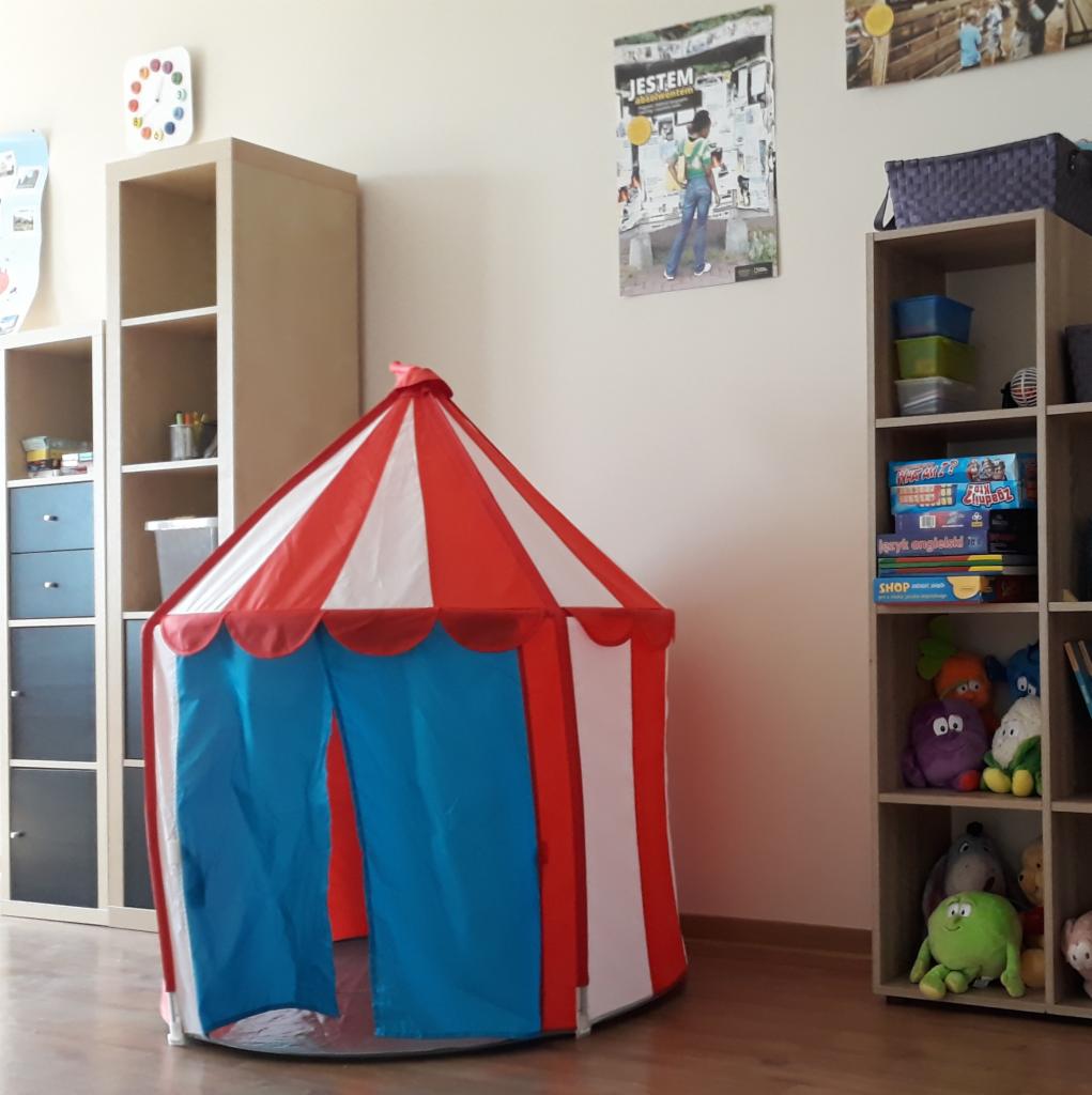 Dziecięcy pokój z namiotem w kształcie cyrku, regałami z zabawkami i plakatem edukacyjnym na ścianie.