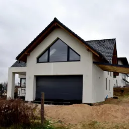 Nowoczesny dom jednorodzinny w trakcie budowy, z widoczną zamontowaną bramą garażową w kolorze antracytowym i oknem trójkątnym na poddaszu, otoczony piaskiem i elementami budowlanymi.