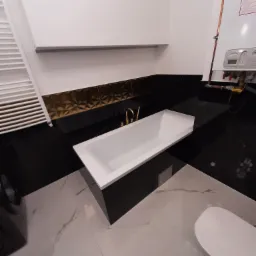 Nowoczesna łazienka z białą wanną w zabudowie, czarnymi płytkami z elementami złota na ścianie i marmurową podłogą, widoczny bojler.