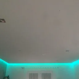 Biały sufit z ukrytym oświetleniem LED w kolorze turkusowym, widoczne okno z firanką w tle