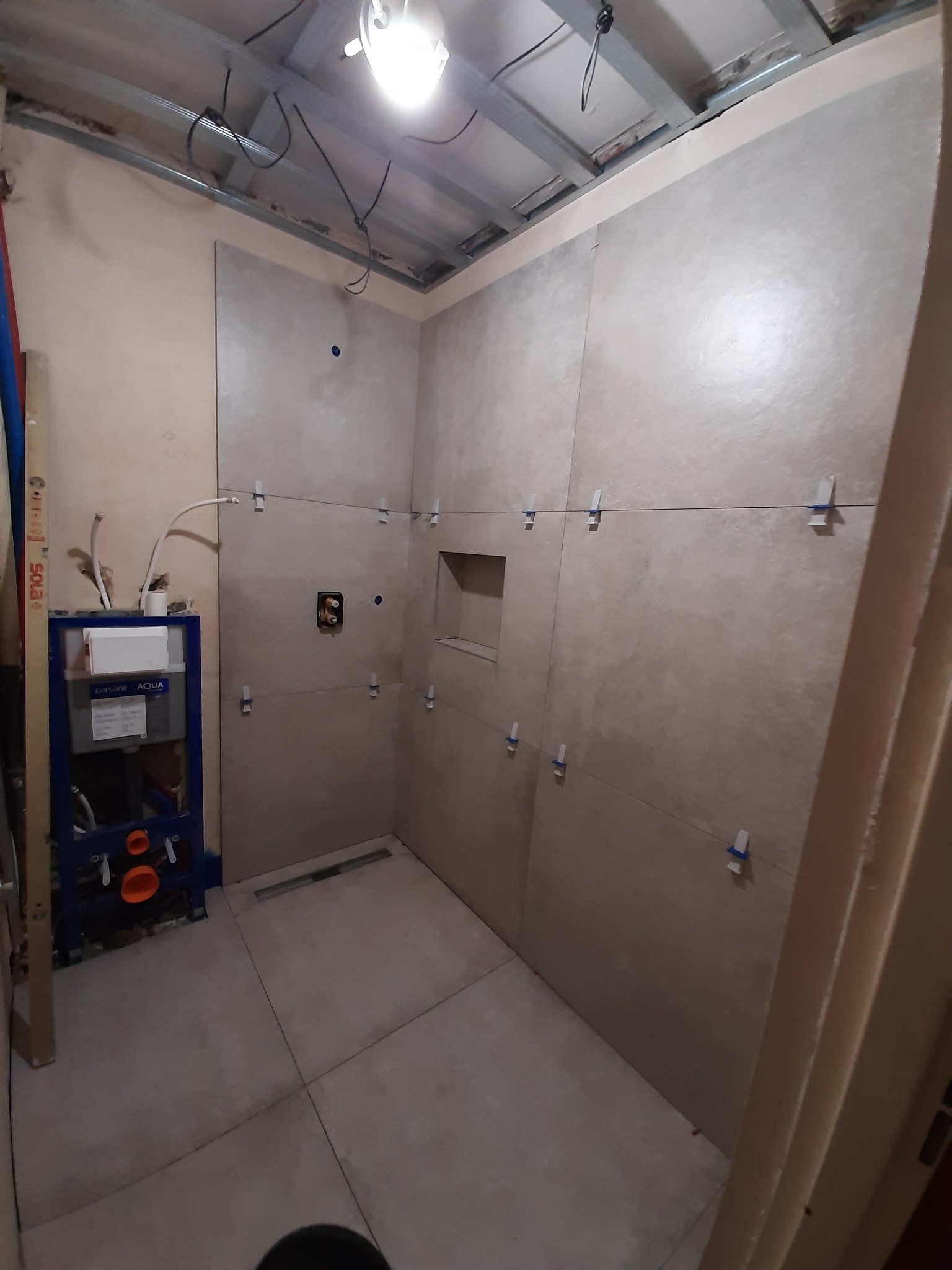 Wnętrze łazienki w trakcie układania płytek ceramicznych, widoczne krzyżyki dystansowe, zabudowa stelaża podtynkowego WC i surowy sufit z metalową konstrukcją.