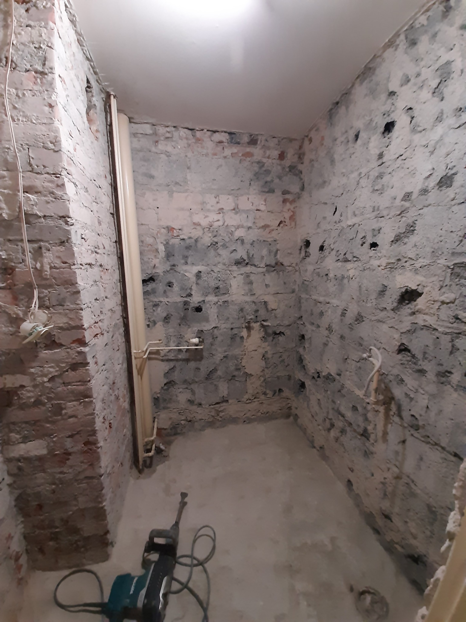 Pomieszczenie łazienkowe w trakcie skuwania starych płytek, widoczne surowe ściany z cegły i betonu oraz narzędzie - młot udarowy.