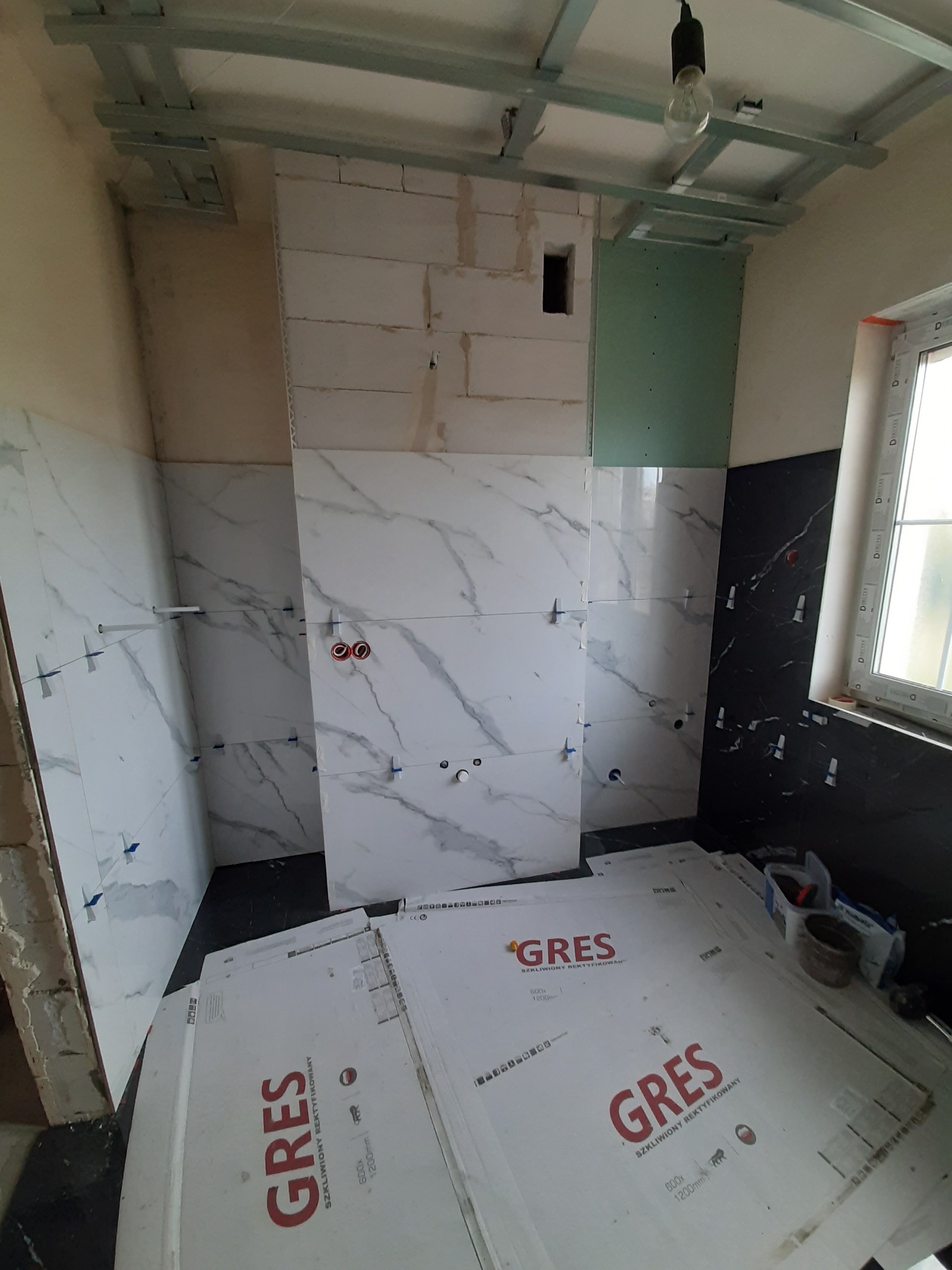 Wnętrze łazienki w trakcie remontu: częściowo ułożone białe płytki imitujące marmur na ścianach, czarne płytki obok okna, płyty kartonowo-gipsowe, widoczne puszki elektryczne, krzyżyki dystansowe...